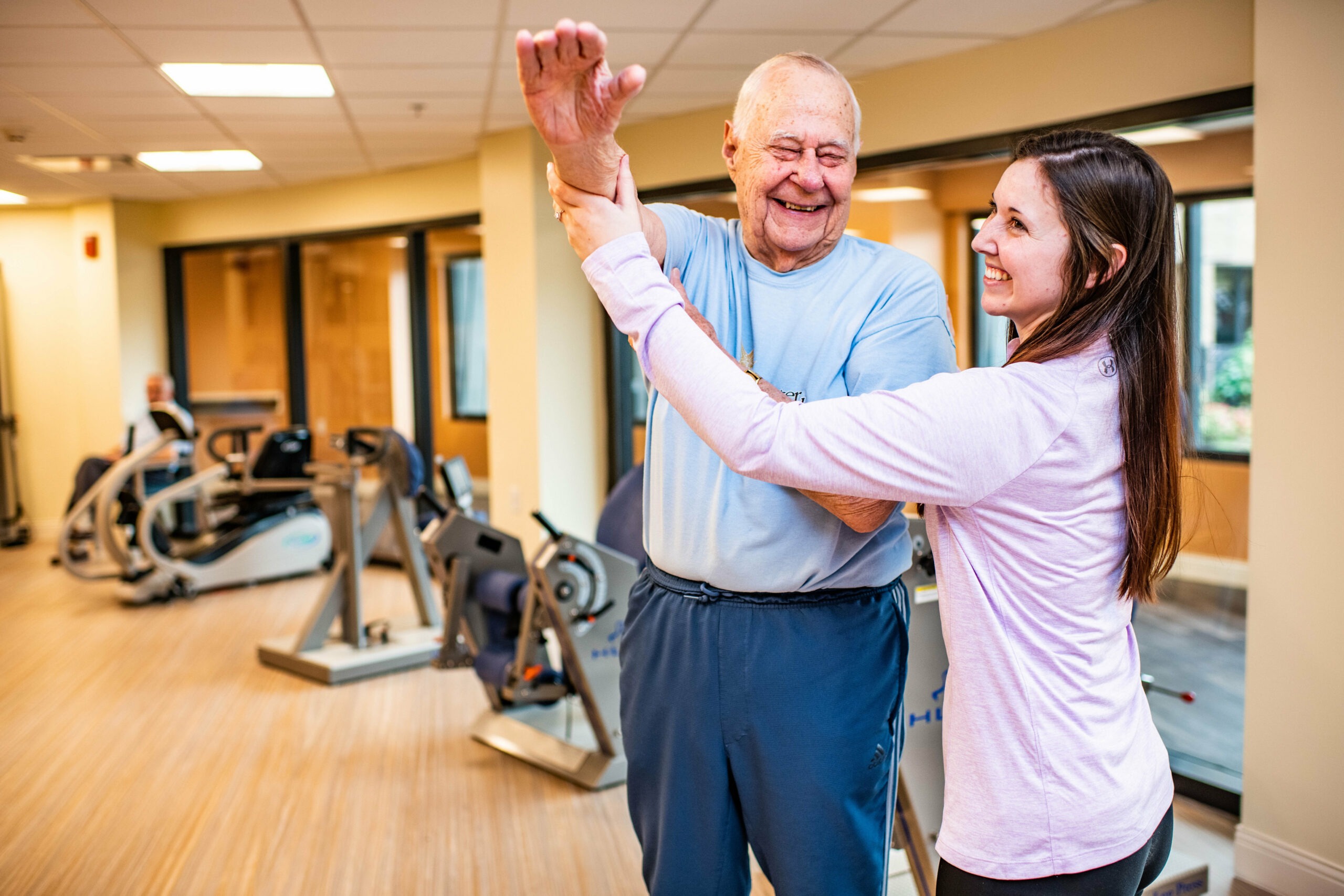 Trainer assisting elderly man with stretches at gym in rehab facility