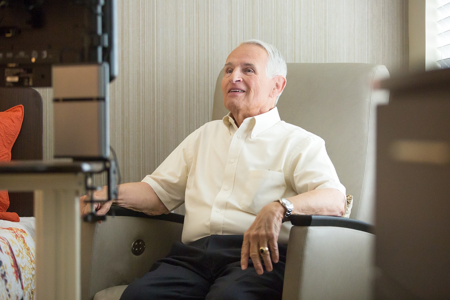 Elderly gentleman sitting in an arm chair smiling a computer screen wearing a light yellow button up shirt