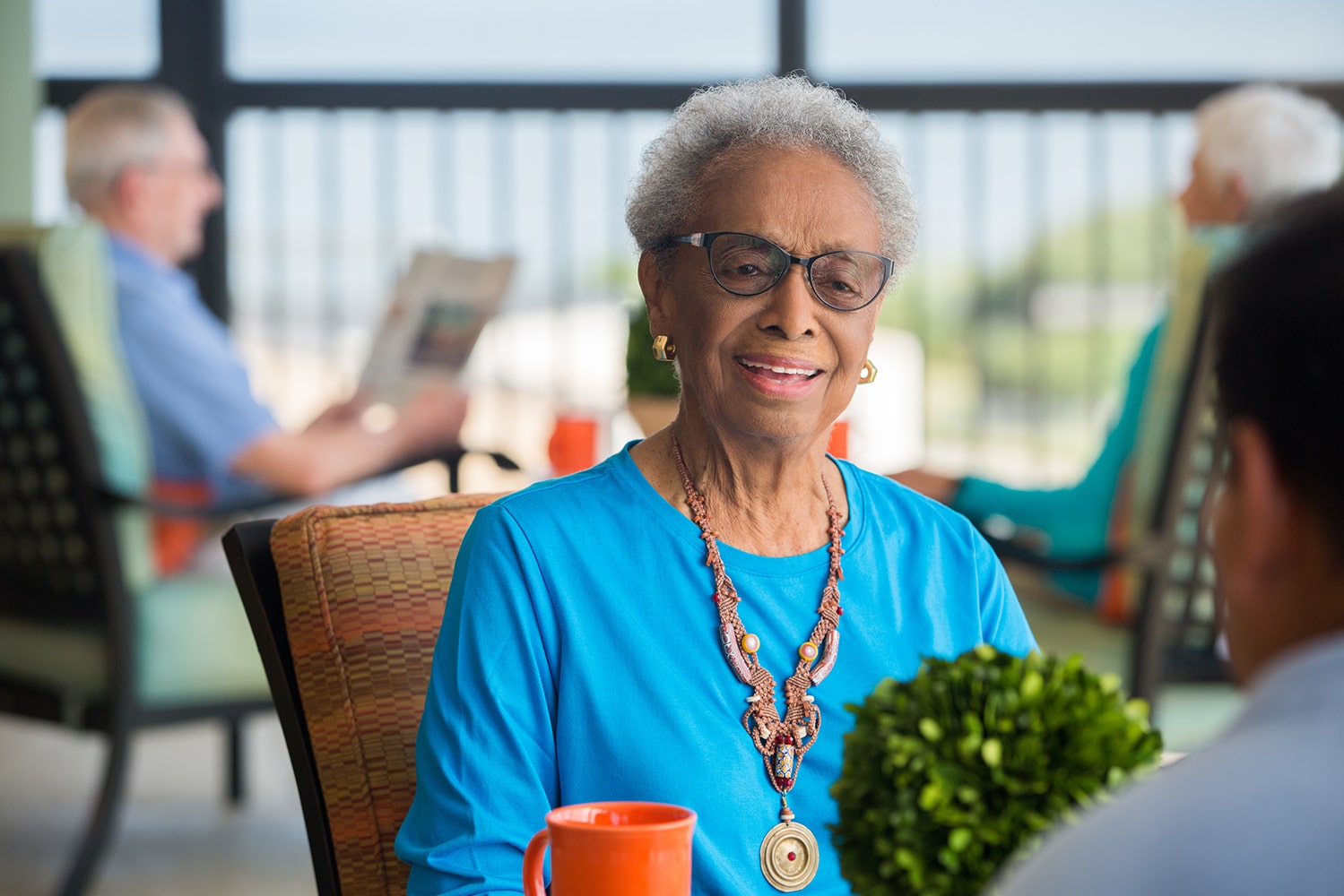 Elderly woman in a blue shirt and large statement necklace on the covered porch smiling at the person she's talking to with an elderly couple sitting in the background reading the newspaper
