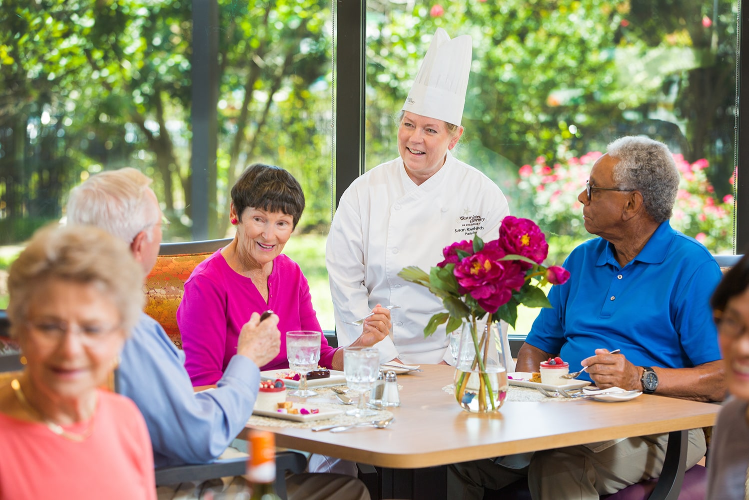 Chef talking to group of people eating dessert at table with vase of pink flowers in the middle