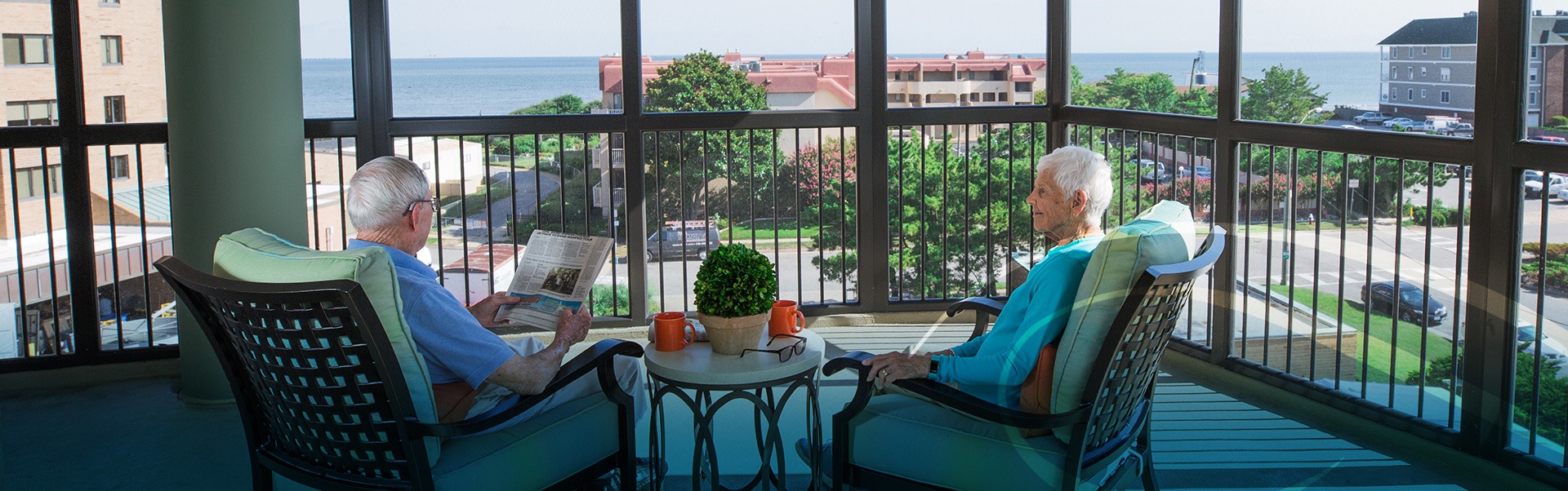 An elderly couple sitting on the covered porch of their residence with one reading the newspaper and the other looking at him read