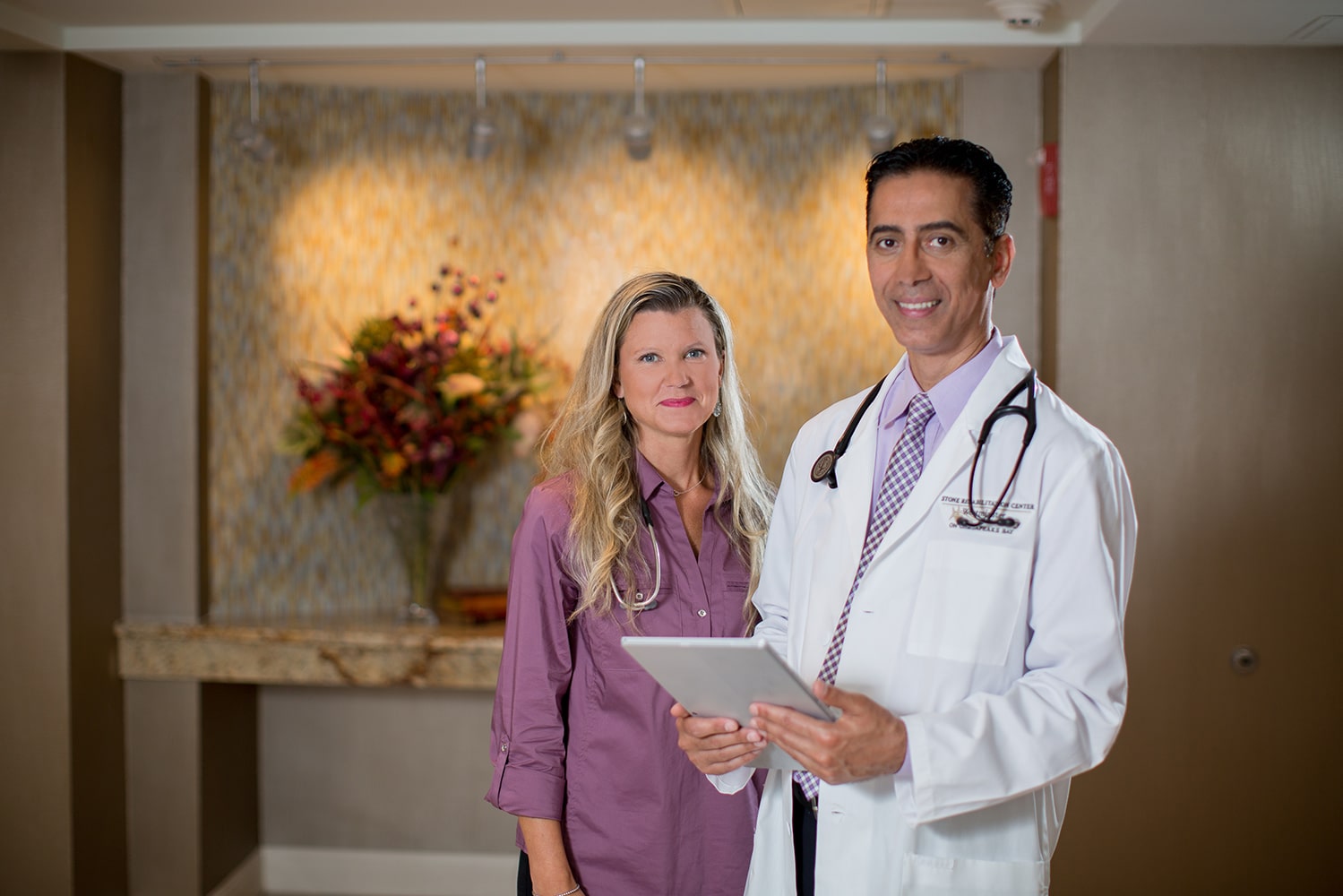 A female and male doctor standing next to each other smiling at the camera wearing stethoscopes around their neck