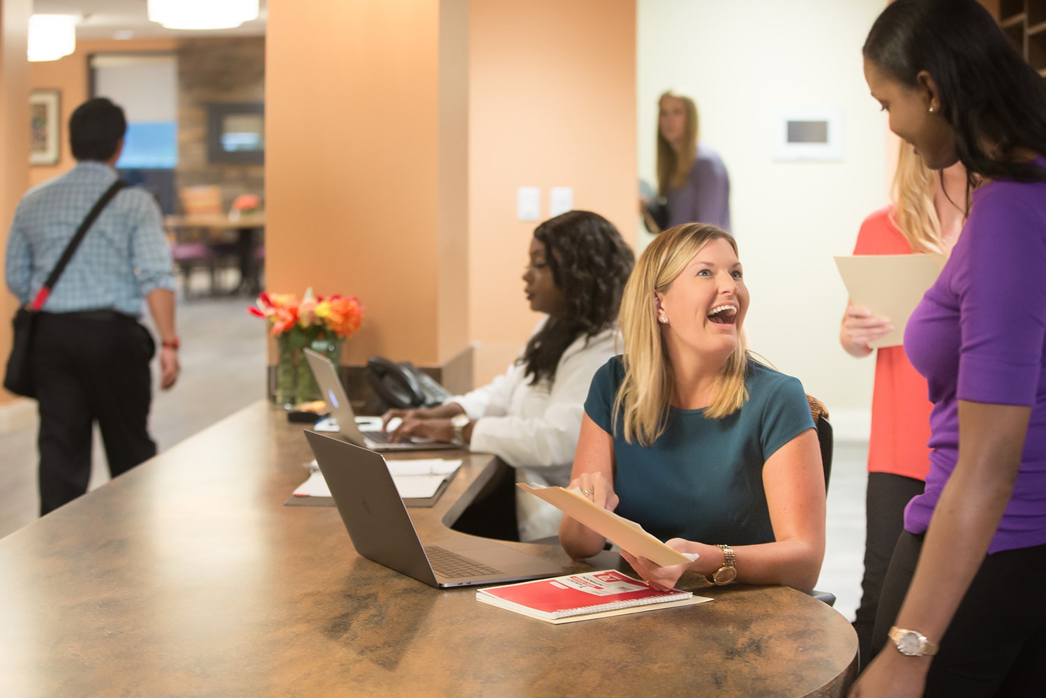 Woman at desk sitting in front of a computer laughing and pointing to a file as she talks to another woman in a purple shirt while various individuals work in the background