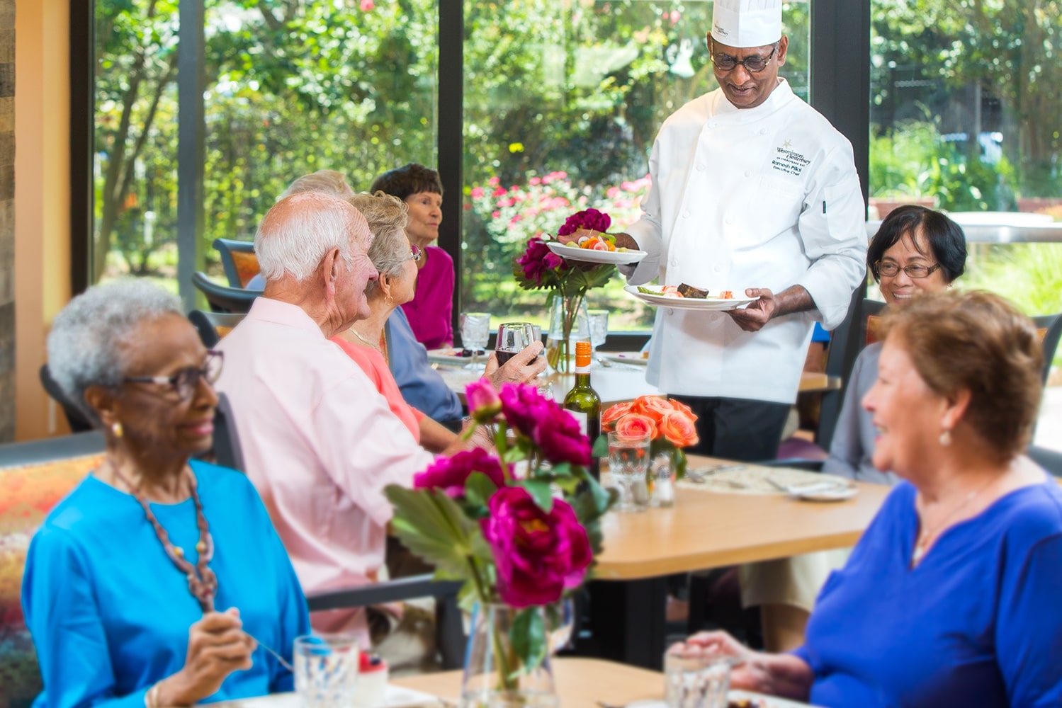 Chef bringing out plates of food to group of people sitting at dinner table and drinking wine in restaurant