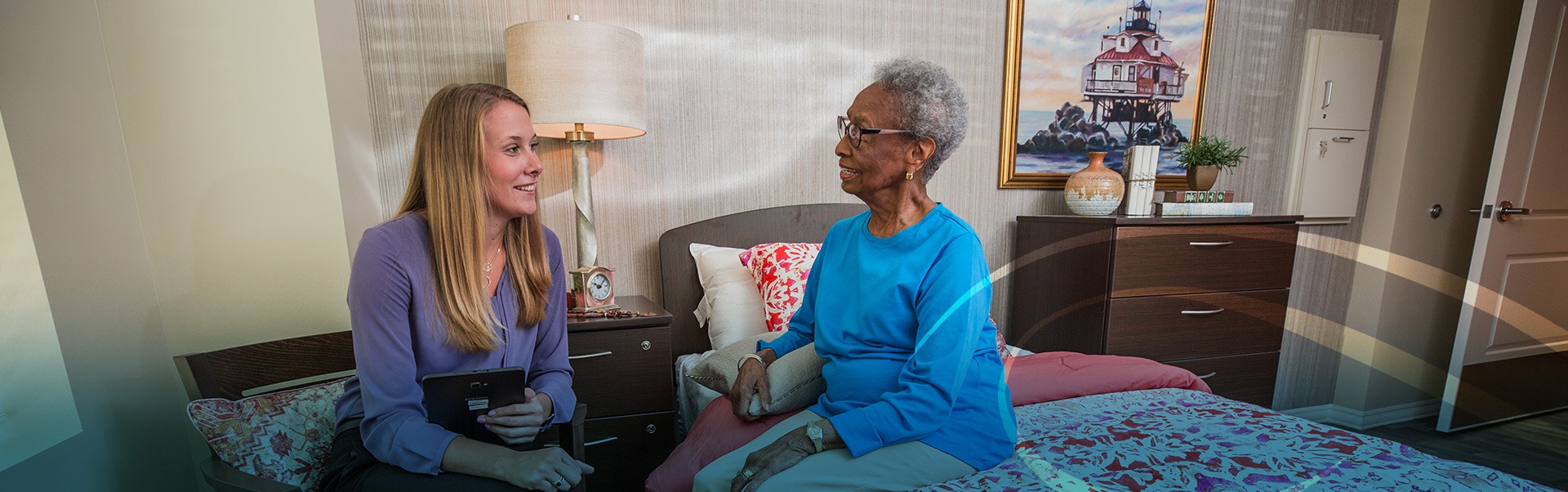 Two women sitting in bedroom at the facility smiling at each other and talking