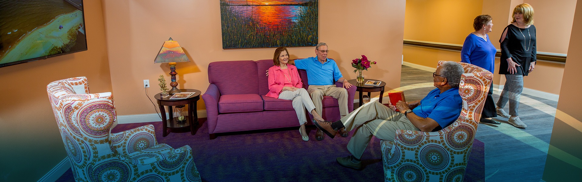 Group of rehab patients sitting together in a sitting room with a colorful couch and chairs while two women walk by in the background