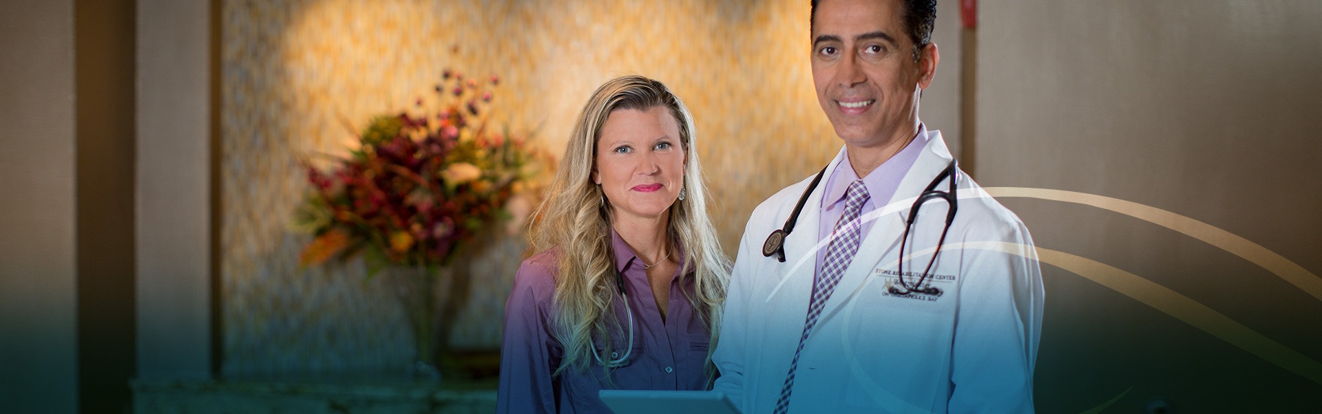 A female and male doctor standing next to each other smiling at the camera wearing stethoscopes around their neck